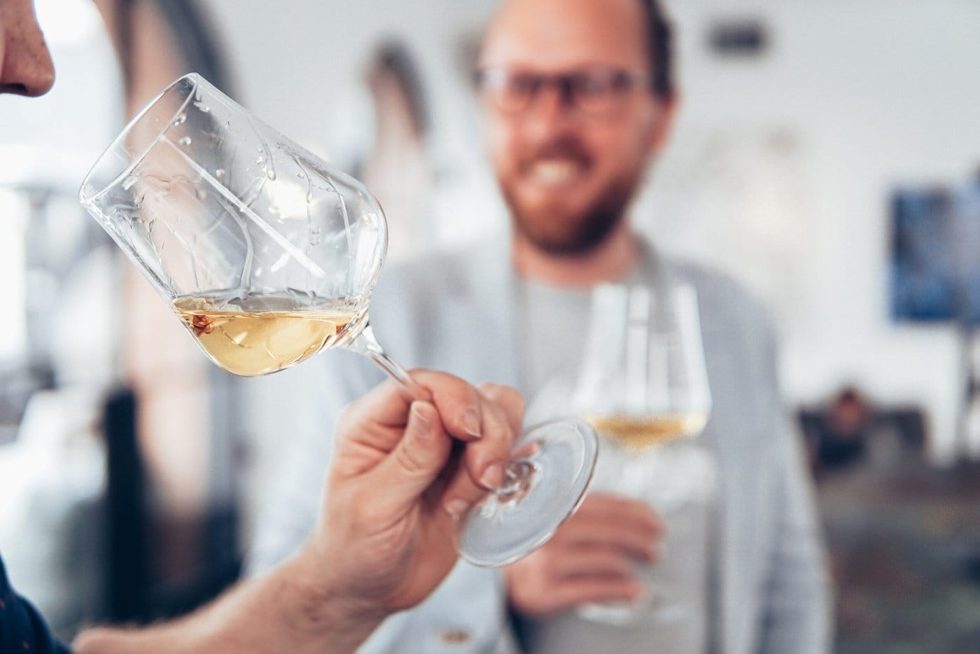 Person holding a glass of wine with man in the background in white shirt