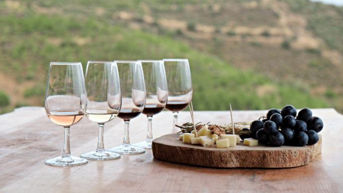 A table with glasses containing types of wines, next to them is a plate of fruit and cheese.