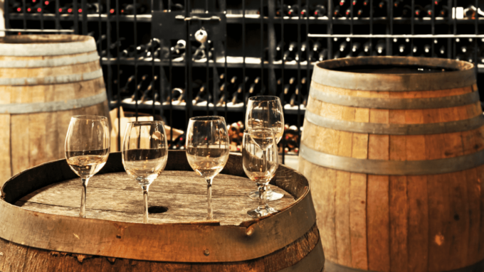 In a wine cellar, rows of wine rest on a stand, in front of it is a barrel with wine glasses.